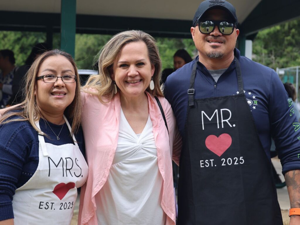 Tarra smiling with two community members wearing Mr. and Mrs est. 2025 aprons at a gathering